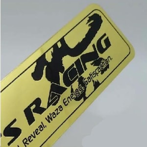 Peut inclure: Un autocollant en métal de couleur dorée avec une silhouette noire d'une personne faisant une pose de course. Le texte "S RACING" est en noir et le texte "Reveal, Waza Energy, Satisfaction." est également en noir.