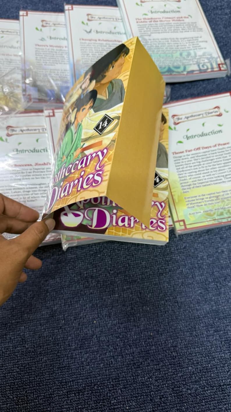 May include: A manga volume titled The Apothecary Diaries volume 14 with a gold-colored cover featuring an anime-style illustration of two characters. The book is partially open, revealing the front cover art and the spine against a background of other books.