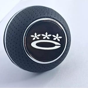 May include: Black leather gear shift knob with a silver trim and a white logo featuring three stylized flowers and a curved line.