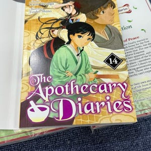 May include: The Apothecary Diaries volume 14 manga book featuring an illustrated cover with two characters in traditional attire. The title is printed in large purple text with a white mortar and pestle icon. The background shows a golden pattern.