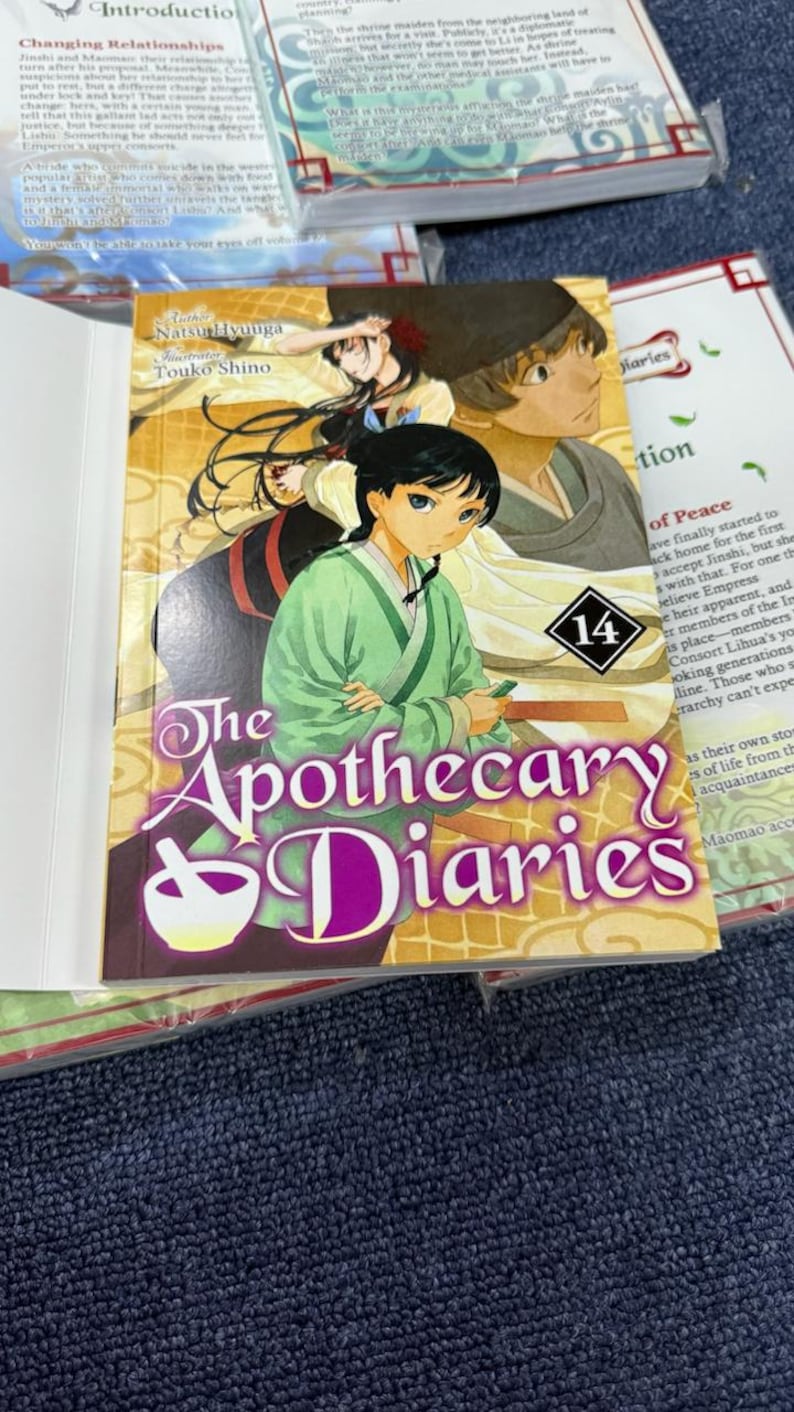 May include: The Apothecary Diaries volume 14 manga book featuring an illustrated cover with two characters in traditional attire. The title is printed in large purple text with a white mortar and pestle icon. The background shows a golden pattern.