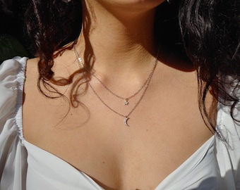 May include: A silver chain necklace with a small star and crescent moon pendant. The necklace is layered and worn by a person with long dark hair.