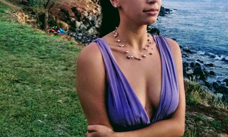 以下が含まれることがあります： 深いVネックの紫色のノースリーブドレスを着た女性。彼女は、重ね着のデザインの繊細な真珠のネックレスをしています。女性は水辺を背景に屋外に立っています。