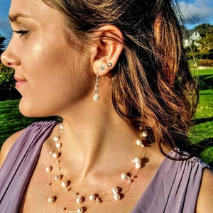 以下が含まれることがあります： 薄い紫色のドレスを着た女性が、繊細な真珠のネックレスとイヤリングをしています。ネックレスは、細いワイヤーに複数の真珠の糸が通され、滝のように連なっています。イヤリングはシンプルで、銀色のフックから真珠が1つぶら下がっています。