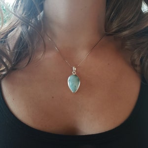 May include: A teardrop-shaped pendant necklace with a light blue gemstone set in a silver-colored metal. The pendant hangs from a delicate silver chain. The necklace is worn against a black top.