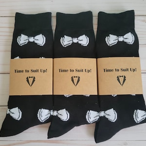 May include: Three pairs of black socks with white bow tie designs. Each pair has a brown paper tag that reads "Time to Suit Up!" with a tuxedo graphic.