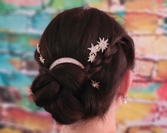 May include: A woman with dark brown hair styled in a bun with a braid. She is wearing several gold star-shaped hair clips and a crescent moon hair clip with rhinestones.