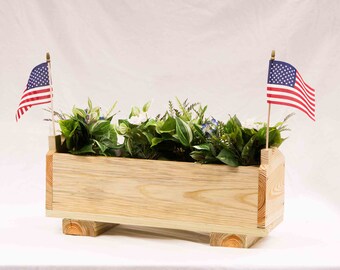 May include: A wooden planter box with a natural finish, featuring two small American flags and a variety of green plants with white and blue flowers.