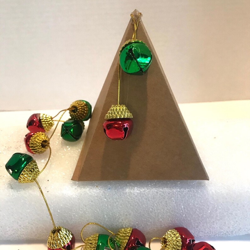 May include: A brown cardboard pyramid with a string of red and green jingle bells hanging from the top. The bells are gold-trimmed and have a shiny finish.