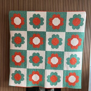 May include: A white quilt with a green and orange floral pattern. The quilt is made up of squares with a flower design in each square. The flowers are in shades of orange and white with a green background.