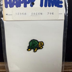May include: A small, green turtle patch with yellow accents, packaged on a white card. The card is labeled "HAPPY TIME" in blue bubble letters. The word "GREEN" is printed on the card.
