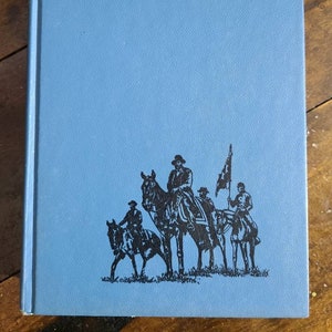 May include: A blue hardcover book with a black silhouette of four people on horseback riding in a line. The people are wearing hats and the person in the front is holding a flag.