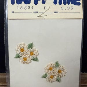 May include: Two embroidered flower patches with cream-colored petals and yellow centers, accompanied by green leaves. The patches are on a white card, packaged in a clear plastic sleeve. The card has the text "HAPPY TIME" in blue.