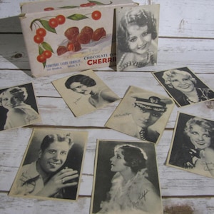 May include: A vintage box of chocolate covered cherries with a collection of signed celebrity trading cards. The box is white with red cherries and the text "CHOCOLATE COVERED CHERRIES" and "VESTROS CANDY COMPANY Buffalo, N.Y.". The cards feature black and white photos of the celebrities with their signatures.