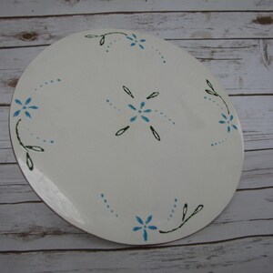 May include: White ceramic plate with a blue and green floral design. The plate has a simple, elegant design with small blue flowers and green leaves.