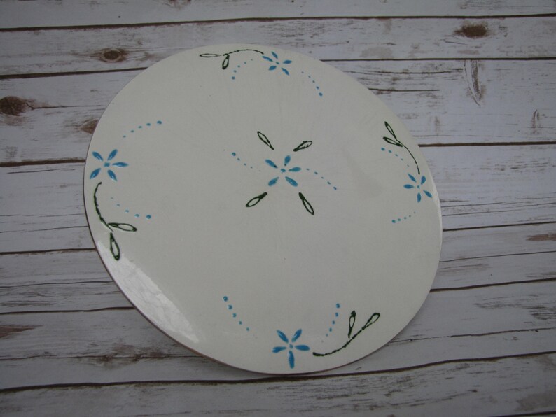 May include: White ceramic plate with a blue and green floral design. The plate has a simple, elegant design with small blue flowers and green leaves.