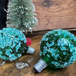 May include: Two green glass light bulbs covered in small, light blue and green glass pieces. The bulbs have a standard screw-in base.
