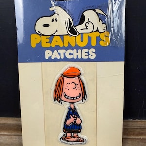 May include: Vintage Peanuts patches on a blue and cream card. The card features Snoopy and the words "PEANUTS PATCHES". One patch shows a smiling cartoon girl with red hair and an orange cap. The patches are sealed in plastic.
