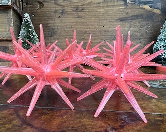 May include: A set of eight pink, sparkly, plastic starburst ornaments. The ornaments have a spiky, star-shaped design and are perfect for adding a touch of vintage Christmas cheer to your holiday decor.