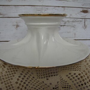 May include: White ceramic cake stand with a gold trim around the top and bottom edges. The stand has a wide base and a tall, narrow center column.