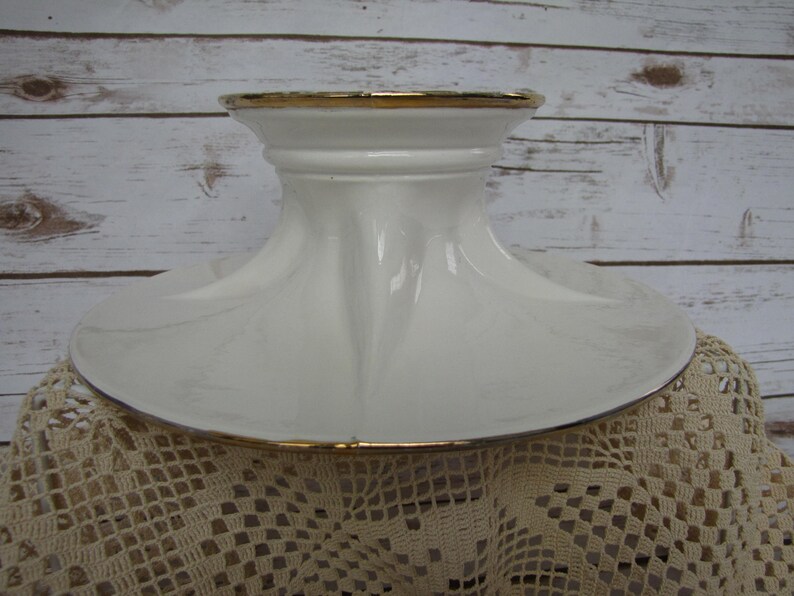 May include: White ceramic cake stand with a gold trim around the top and bottom edges. The stand has a wide base and a tall, narrow center column.