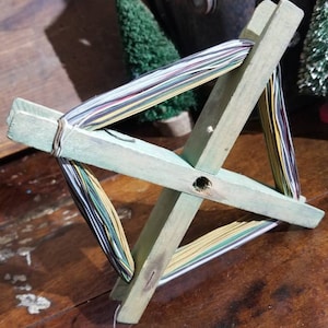 May include: A wooden yarn winder with a green paint finish. The winder has a cross shape and is holding a bundle of colorful yarn.