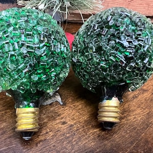 May include: Two green glass light bulbs covered in small green glass pieces. The bulbs have a standard screw-in base.
