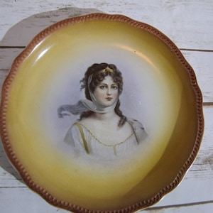 May include: A vintage porcelain plate with a yellow background and a painted image of a woman with a white scarf and a gold headband.