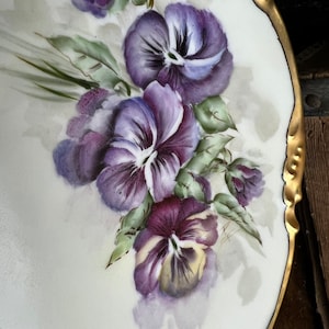May include: A white ceramic plate with a gold rim features a hand-painted design of purple pansies with green leaves. The pansies are painted in a watercolor style, with soft edges and subtle shading.