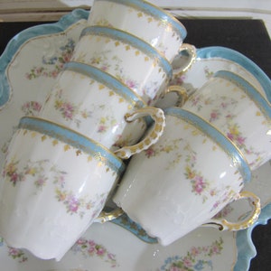 May include: A set of five vintage porcelain teacups with floral patterns and gold accents. The cups are stacked on a blue and white scalloped serving tray.