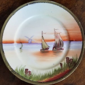 May include: A decorative plate featuring a painted seascape with sailboats, a windmill, and grassy foreground. The plate has a cream-colored center with a gold-colored rim and a painted sunset in the background.