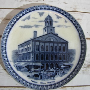 May include: Blue and white ceramic plate with a depiction of Faneuil Hall in Boston, Massachusetts. The plate has a decorative border with a repeating pattern.