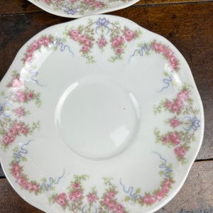 May include: A white porcelain saucer with a scalloped edge features a delicate floral pattern of pink roses and blue ribbons.