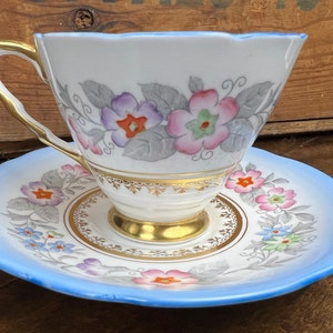 May include: A vintage teacup and saucer set with a floral pattern. The cup and saucer are white with a blue rim and feature a delicate floral design in shades of pink, purple, and green. The cup has a gold trim around the rim and base.