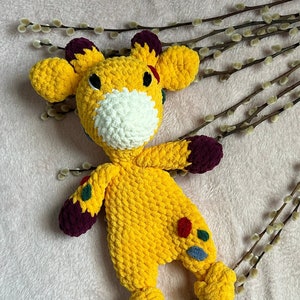 May include: A handmade yellow crochet giraffe stuffed animal with a white muzzle, dark purple hooves, and colorful spots. The giraffe has large ears and is lying on a pink surface.