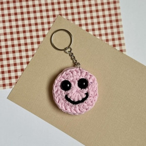 May include: A pink crocheted smiley face keychain with black button eyes and a stitched smile. The keychain is attached to a silver metal ring and chain. The background includes a tan and a red and white checkered paper.