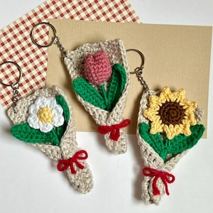 May include: Three crocheted flower bouquets with keychains. The bouquets are wrapped in beige yarn and tied with red ribbon. The flowers are a white daisy, a pink tulip, and a yellow sunflower.