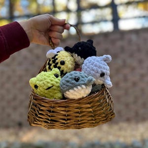 May include: A woven basket filled with handmade crochet plush toys. The basket contains a yellow bee, a black and white whale, a light green frog, a gray shark, and a light blue dolphin. The toys are soft and round, with black safety eyes.