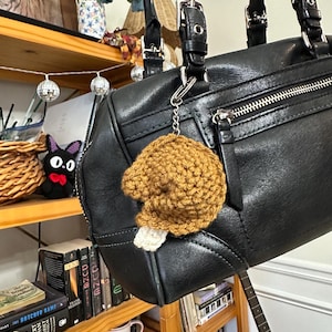 May include: A handmade, crocheted turkey leg keychain in shades of brown and white, hanging from a black leather handbag. The turkey leg is detailed with textured yarn, and the keychain is attached with a silver-colored clasp. The bag has silver zippers and buckles.