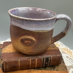 May include: A handmade ceramic mug with a dragonfly emblem. The mug features a brown base, a white and purple rim, and a curved handle. The mug is sitting on top of a stack of old books.