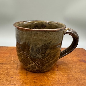 May include: A brown ceramic mug with a fern leaf design. The mug has a rounded body and a handle.