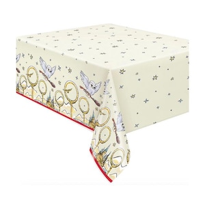 May include: Cream-colored tablecloth featuring a repeating pattern of owls on brooms, golden hoops, and castle illustrations. The tablecloth has a red border and is decorated with small black star shapes.