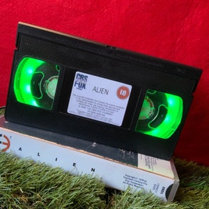 May include: A black VHS tape with a green light shining through the window. The tape is labelled "Alien" and has a CBS/FOX Video logo. The tape is rated 18.