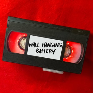 May include: A black VHS tape with red labels and the text "WALL HANGING BATTERY" on a white label.