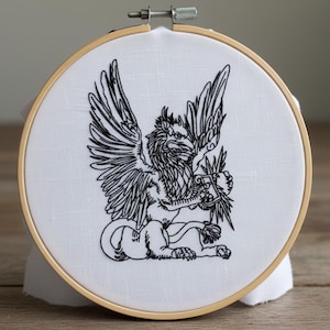 Könnte beinhalten: Gesticktes Greifen-Design auf weißem Stoff, gespannt in einem hölzernen Stickrahmen. Der Greif, ein Fabelwesen, ist in schwarzem Faden mit detaillierten Flügeln und einem Löwenkörper dargestellt. Der Rahmen wird mit einem Metallverschluss befestigt.