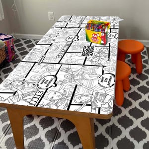 Könnte beinhalten: Ein Holztisch mit Comic-Design, der schwarz-weiße Illustrationen und Text zeigt. Eine Schachtel mit 64 Crayola-Buntstiften steht auf dem Tisch. Zwei kleine orangefarbene Hocker stehen neben dem Tisch.