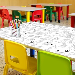 May include: A white table with a black and white princess-themed colouring table cloth. There are coloured pencils on the table and a yellow chair in the foreground.