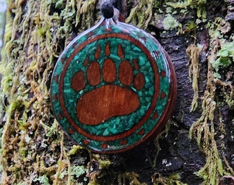 Puede incluir: Un colgante redondo de madera con un diseño de huella de pata de oso verde y marrón. El colgante está en un cordón negro y está colgando de una rama de árbol.
