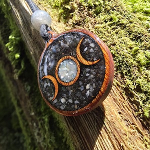 May include: A circular pendant featuring a dark resin inlay filled with black stone fragments and a central white crystal. The piece is framed in polished wood and displays a triple moon symbol in wood grain. It hangs from a black cord with a bead.
