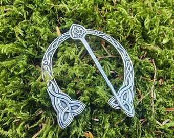 May include: A silver Celtic knot brooch with a decorative design. The brooch is in the shape of a circle with a pin in the center. The brooch is on a bed of green moss.